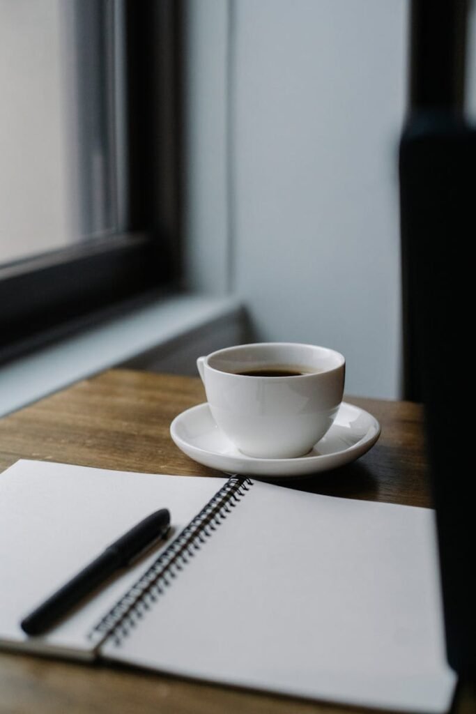 A cup of coffee and an open notebook by a window in a cozy café setting.